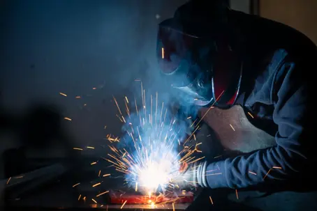 Welder in protective gear skillfully joining metal pieces, generating a dazzling display of sparks and smoke in a dimly lit workshop filled with industrial machinery